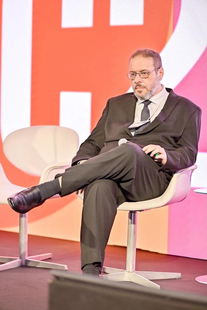 Marco Stephan Lofrano Alves sentado numa poltrona branca no palco de um debate, de pernas cruzadas e microfone na mão.
