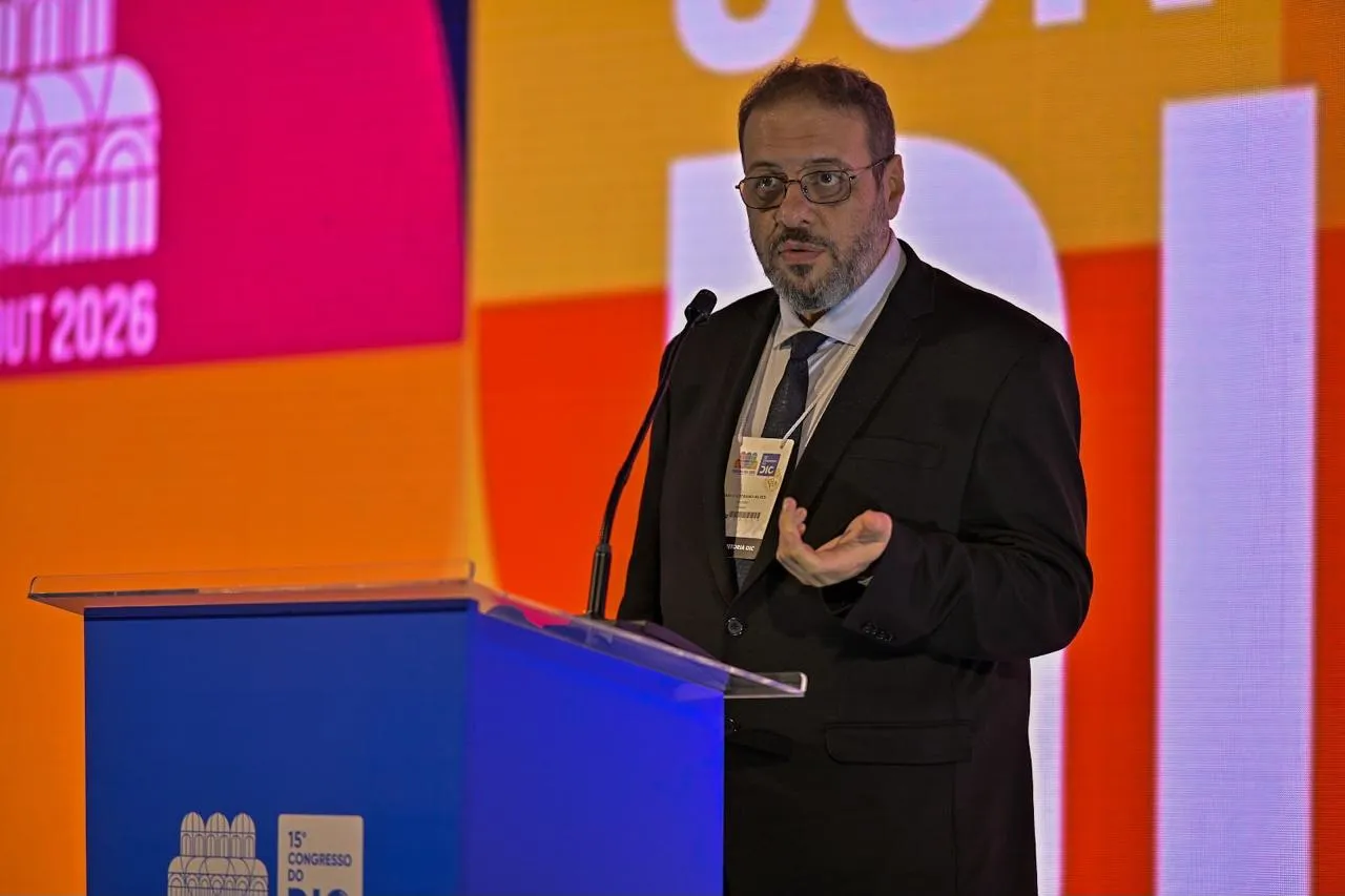 Marco Stephan Lofrano Alves fala ao microfone num púlpito, gesticulando, diante do painel colorido do congresso.