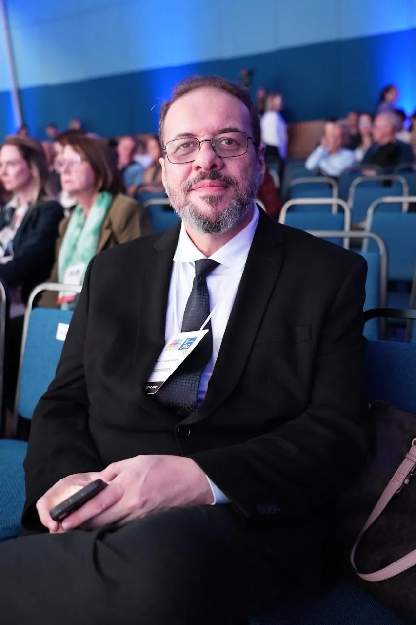 Marco Stephan Lofrano Alves sentado na plateia do 15º Congresso do DIC, de terno e crachá do evento.
